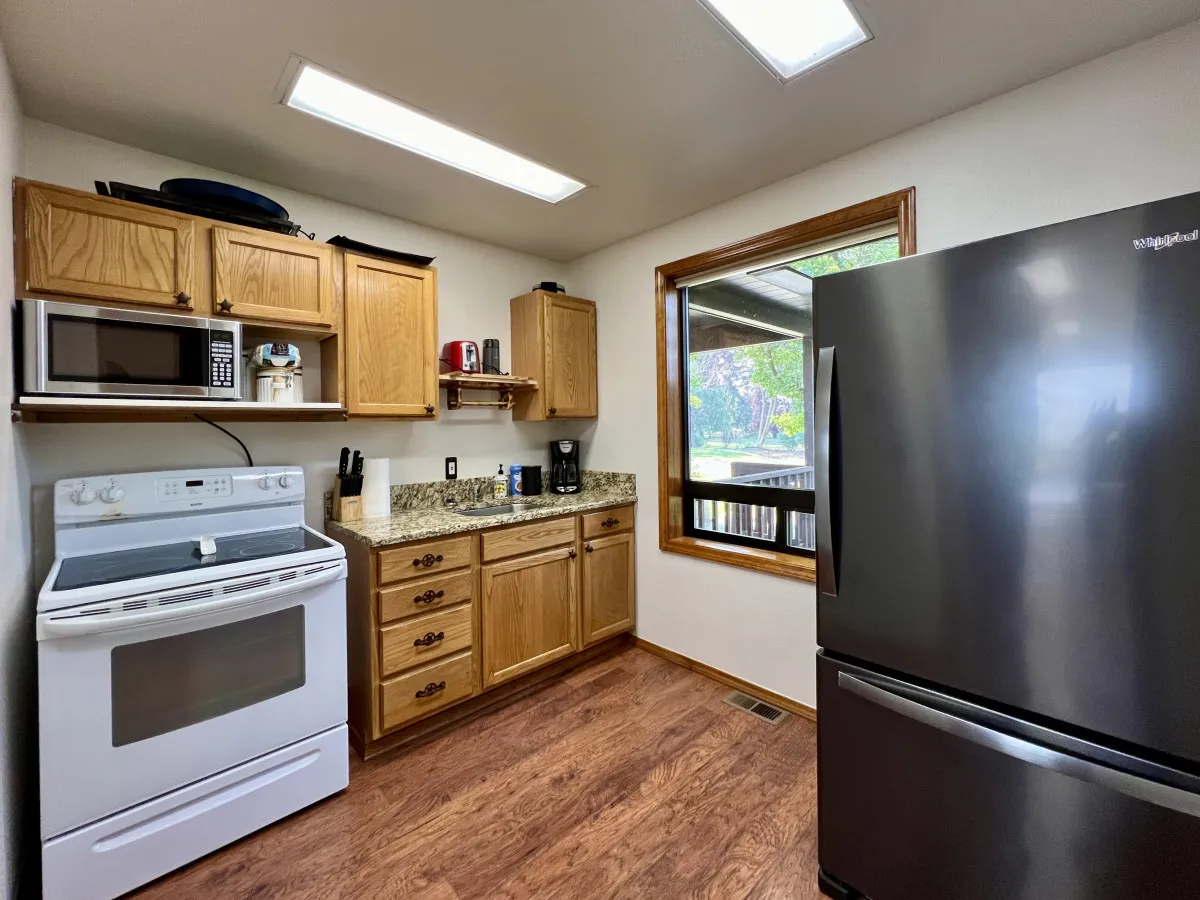 A welcoming kitchen area featuring a stainless steel refrigerator, granite countertops, warm wooden cabinets, and a window that lets in natural light. The scene suggests a place where family meals and morning coffee happen naturally.