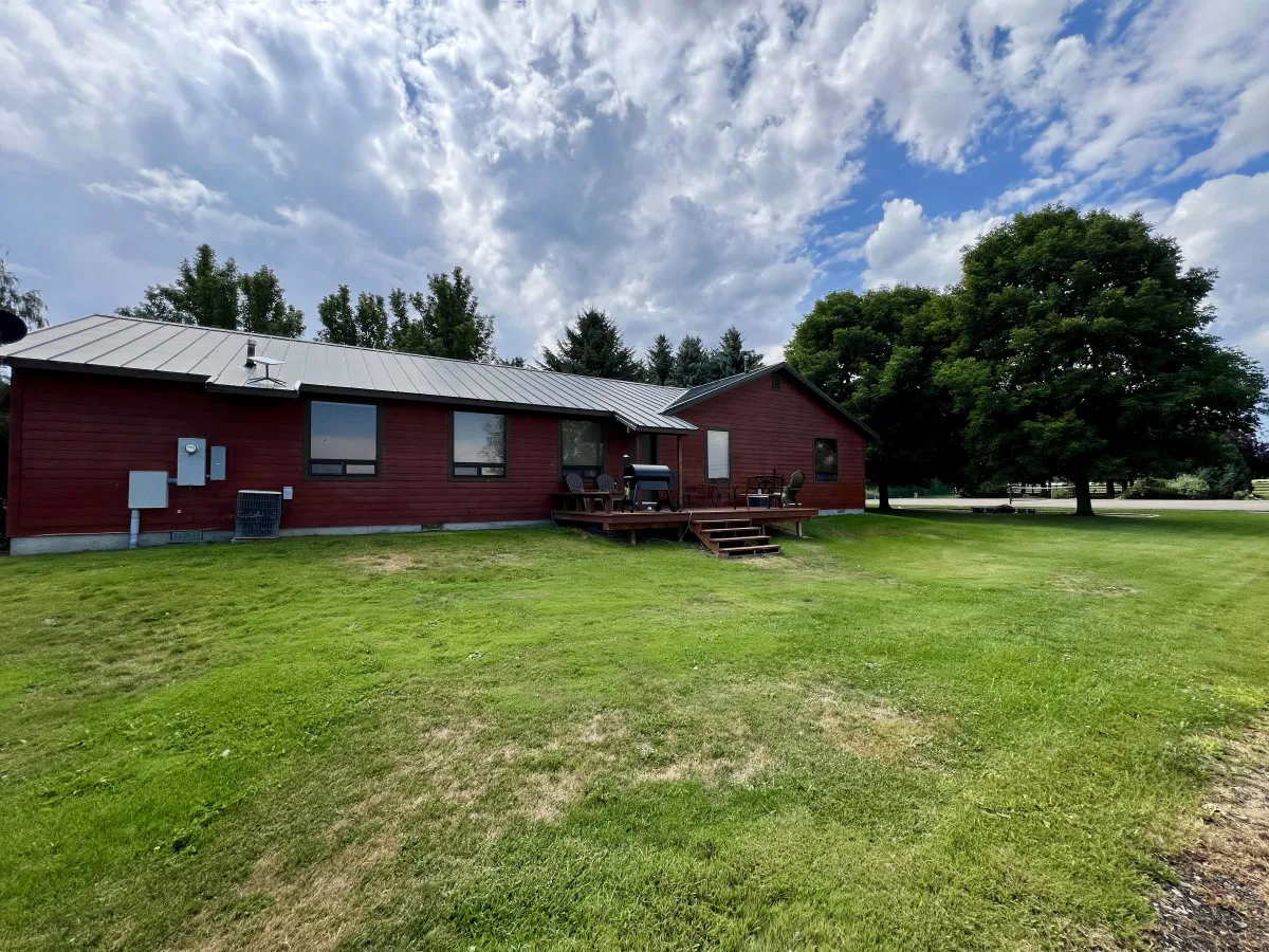 A welcoming red wooden house features a back deck with seating overlooking a large green lawn. A mature tree offers shade nearby, and the open lawn extends into the distance—ideal for gatherings and outdoor activities.