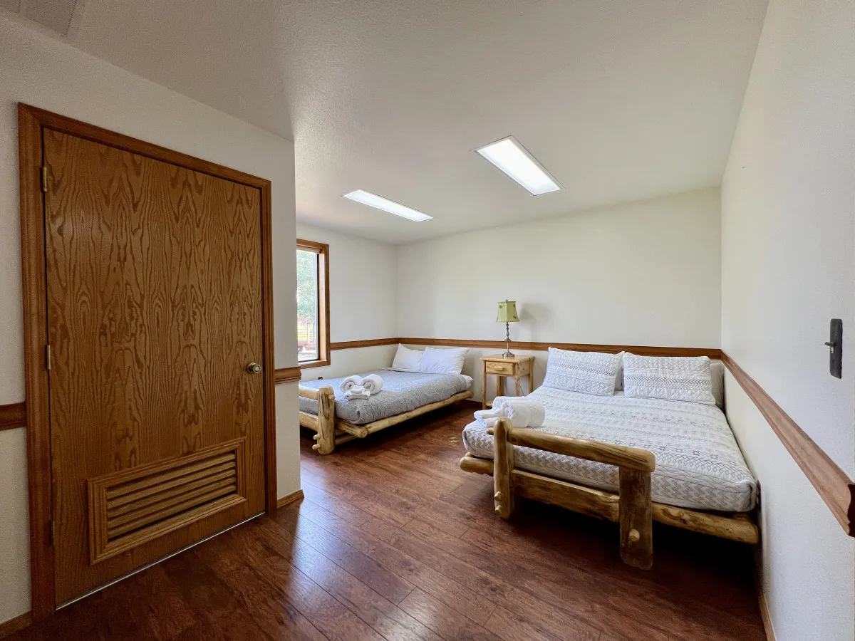 A bright bedroom with two log-frame beds, a window allowing ample natural light, and a nightstand topped with a lamp. The arrangement suggests shared moments for families or friends, with a cozy, welcoming vibe that encourages togetherness.