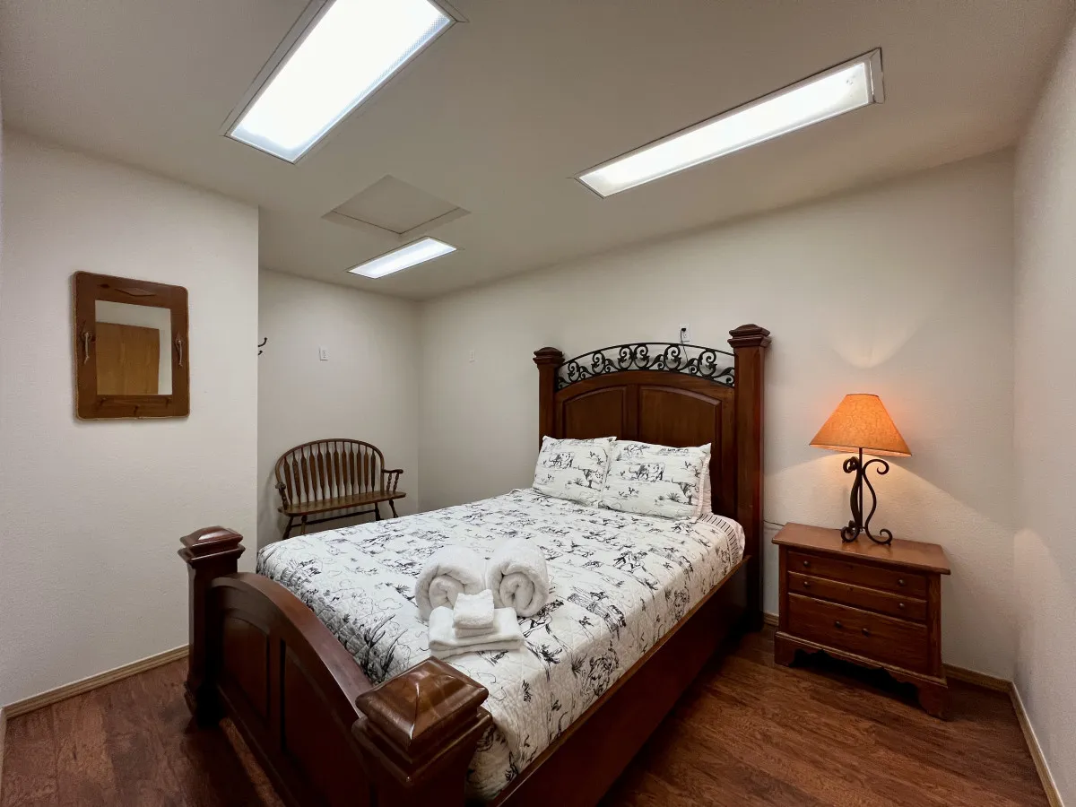 A warmly lit bedroom featuring a classic wooden bed with an ornate headboard, a decorative lamp casting a soft glow, and a cozy upholstered chair perfect for reading or relaxing. The space feels intimate, inviting, and ideal for unwinding after a busy day.
