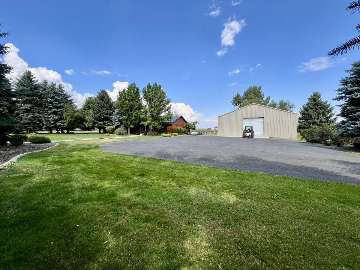A sprawling green lawn stretches across the backyard, bordered by a tree line and a well-kept pathway. A metal storage building sits in the background, suggesting ample space for play and utility. The vast open area is ideal for family games, picnics, and outdoor gatherings.
