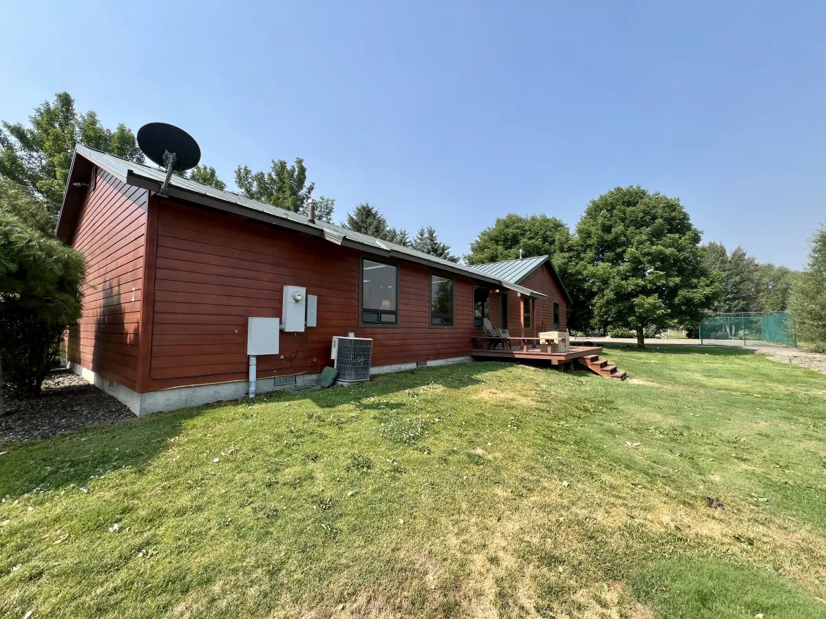 A red wooden deck with comfortable seating overlooks a green lawn and is framed by large mature trees. The setup implies a casual, inviting space for family gatherings or a peaceful moment outdoors.