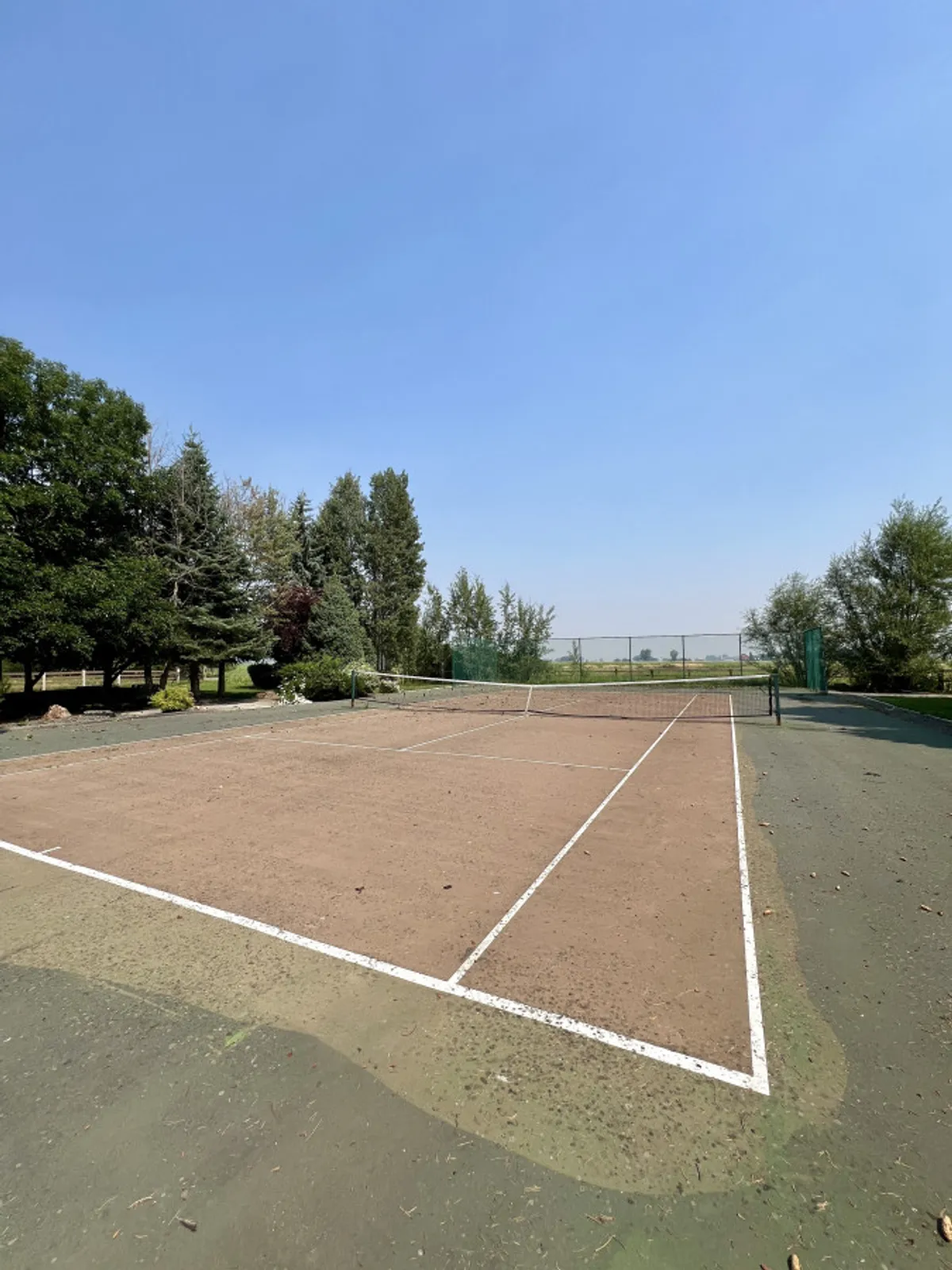 A well-maintained tennis court surrounded by leafy trees, with the net ready for a match. The environment feels energetic yet serene, inviting residents and guests to play or spectate.
