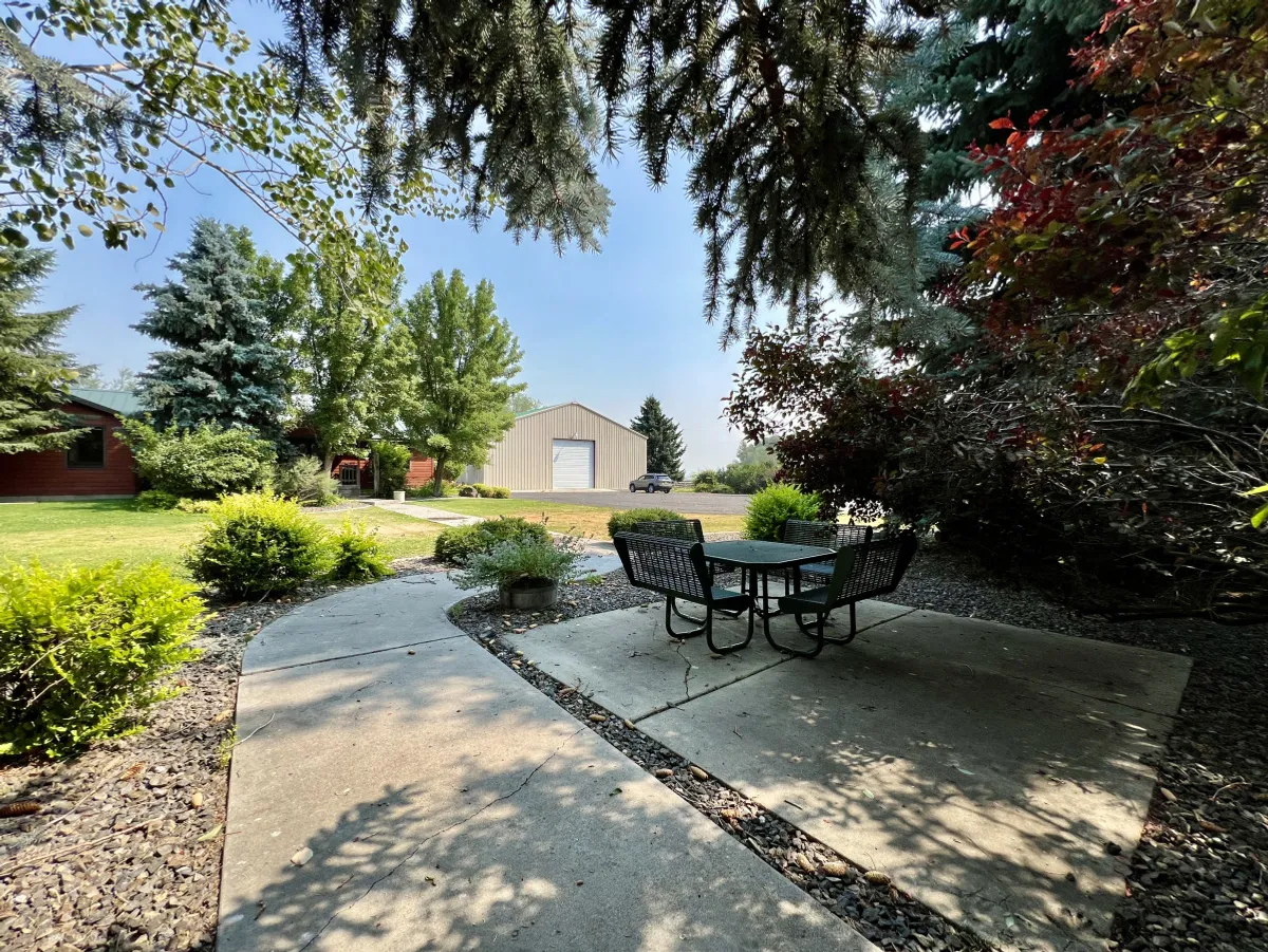 This exterior scene features a patio table with chairs set amid lush greenery, with a distant barn visible. The setting is ideal for outdoor dining, family gatherings, and unwinding after activities like a round of golf.