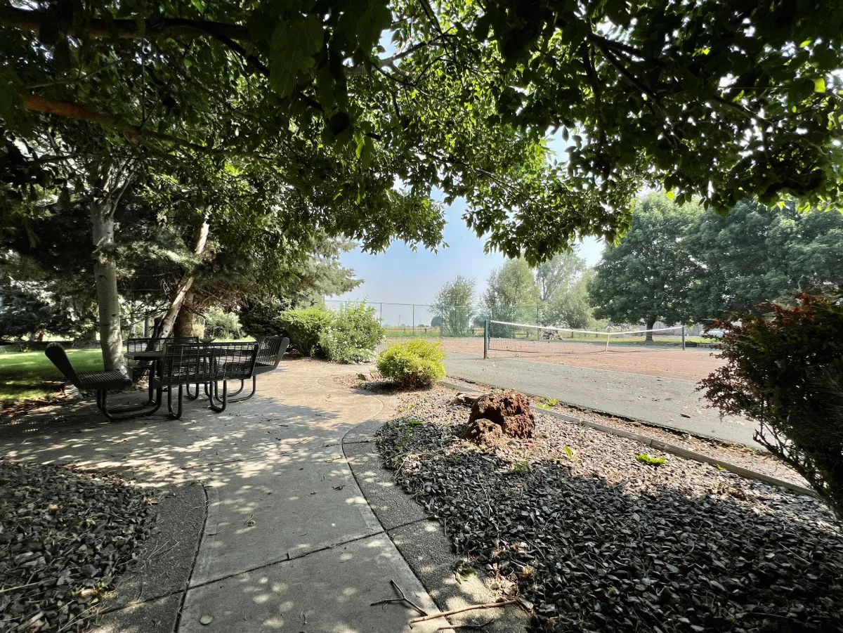 An exterior view featuring an outdoor dining table set among lush greenery, with a pathway and the tennis court visible in the background. The scene evokes a peaceful atmosphere for group gatherings and outdoor meals.