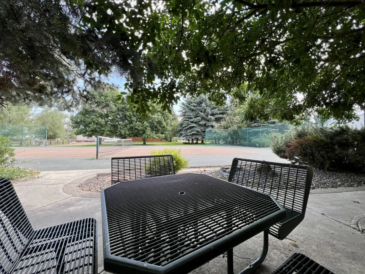 This image shows a metal outdoor table with surrounding trees and the tennis court visible in the background. The setting is tranquil, suggesting a place to relax, socialize, or enjoy refreshments after playing.
