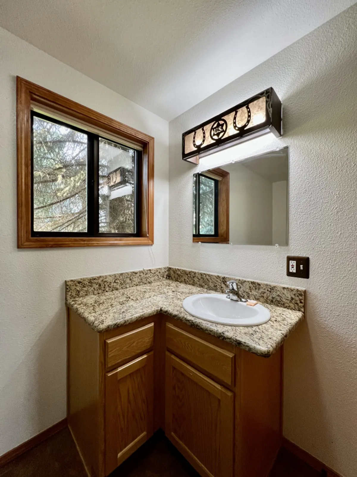 A bathroom with a granite countertop, a well-lit mirror, a sink, and a window framing a tranquil natural view. The scene exudes a sense of calm and practicality, offering a serene atmosphere for morning routines or evening wind-downs.