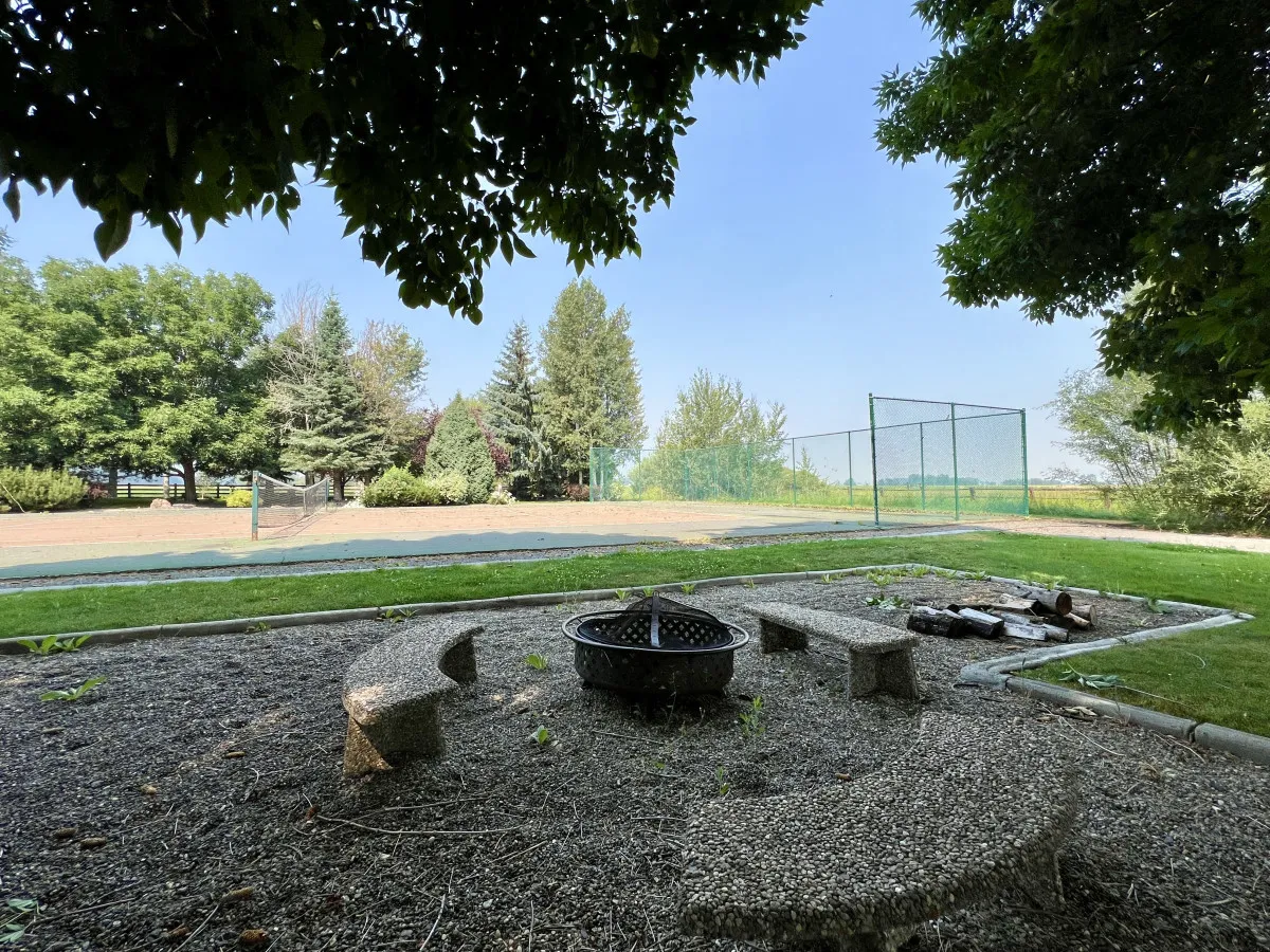 The image showcases a cozy fire pit area with surrounding seating, lush greenery, and a tennis court visible in the background. The communal setup emphasizes outdoor gatherings, storytelling, and activities in a serene mountain environment.